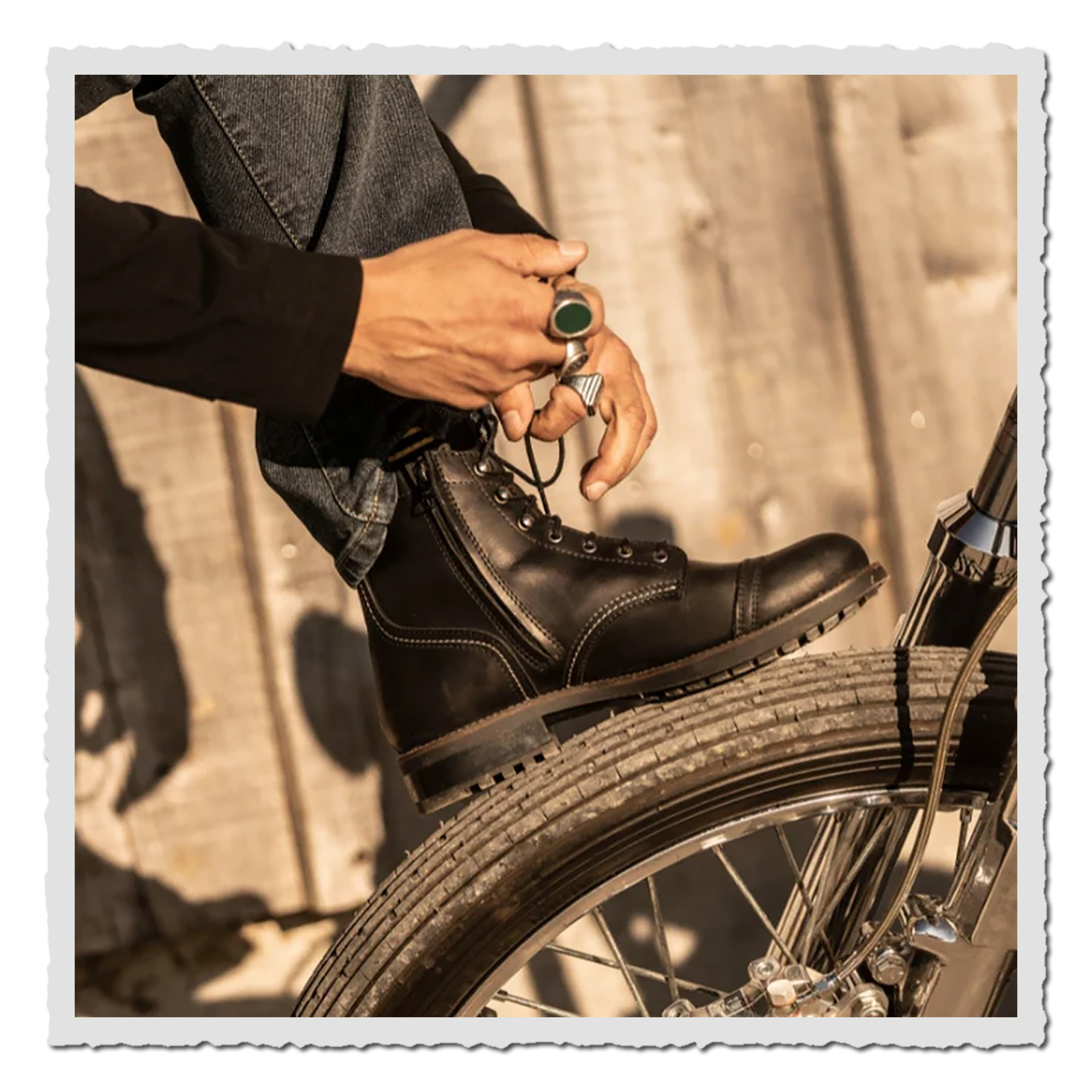 Schwarzer Schnürstiefel auf Vorderrad eines Motorrads, Hand mit Ringen, warmes Sonnenlicht, Holz im Hintergrund.